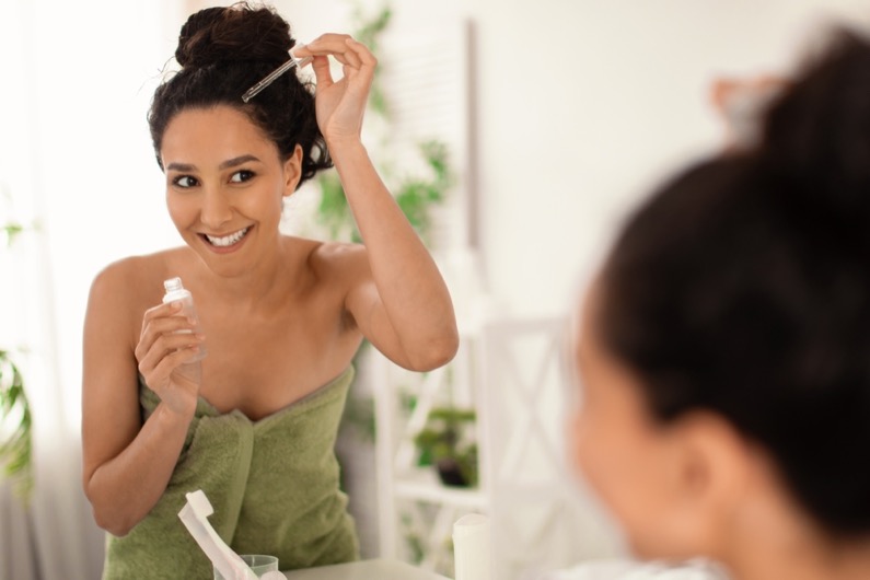 Cheery young woman in towel applying serum or natural oil on her scalp near mirror at home, copy space. Organic cosmetics for haircare, damaged dry hair treatment, daily beauty routine concept